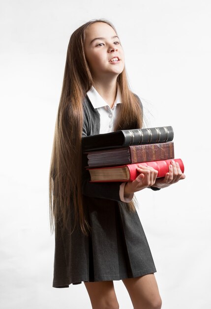 Retrato de colegiala llevando una pesada pila de libros contra el fondo blanco.