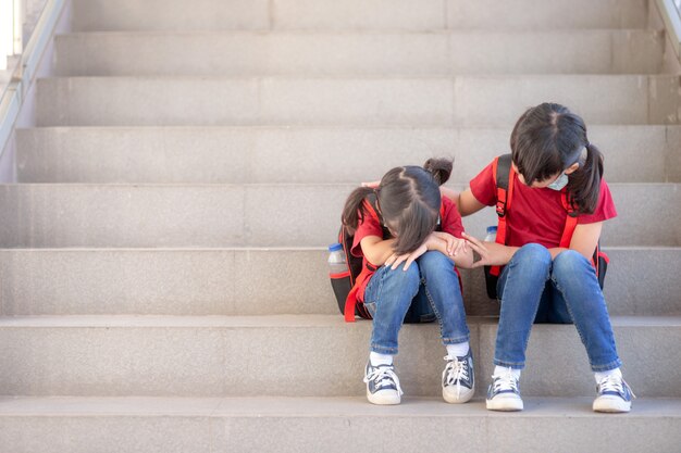 Retrato de colegiala llorando mientras está sentado en las escaleras al aire libre con sonriente hermana consolándola, espacio de copia