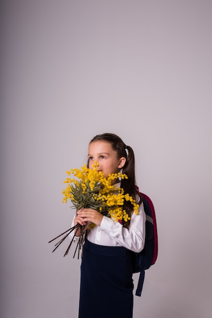 Retrato de una colegiala morena en uniforme con una mochila se encuentra con un ramo de mimosa sobre un fondo blanco con un lugar para el texto