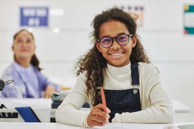 Retrato de una colegiala negra sonriente con gafas sentada en el escritorio en el aula y mirando la cámara