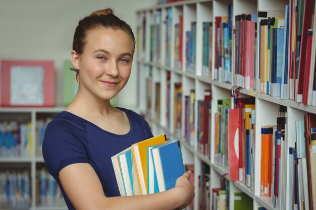 Retrato de colegiala de pie con pila de libros en la biblioteca