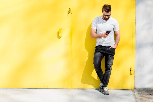 Retrato colorido de um homem bonito vestido de camiseta branca e jeans com telefone no fundo amarelo