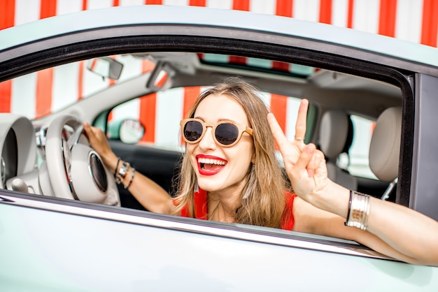 Retrato colorido de uma jovem feliz dirigindo um carro no fundo da parede vermelha durante as férias de verão