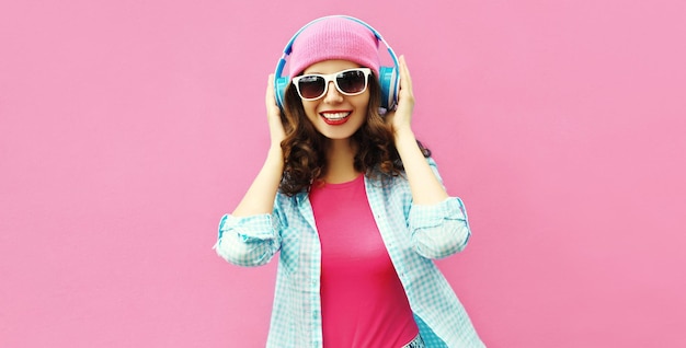 Retrato colorido de una joven feliz escuchando música con auriculares y sombrero sobre un fondo rosa