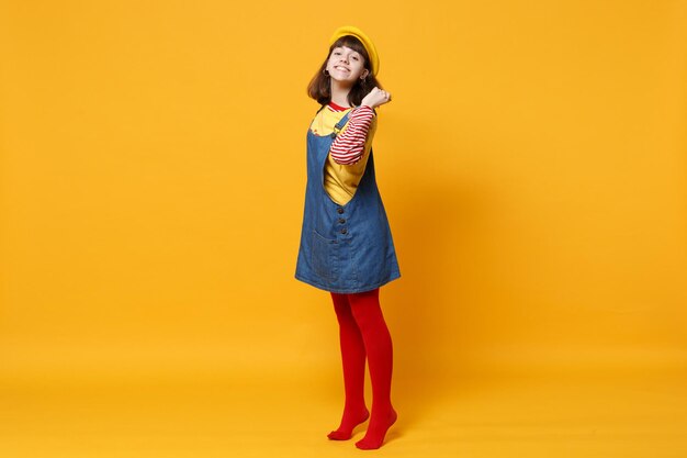 Retrato completo de una adolescente bonita con boina francesa, vestido de mezclilla sosteniendo el pelo aislado en el fondo de la pared amarilla en el estudio. Emociones sinceras de la gente, concepto de estilo de vida. Simulacros de espacio de copia.