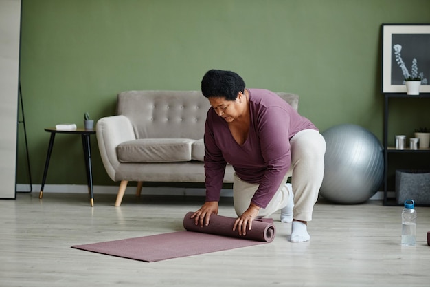 Retrato completo de una anciana negra desenrollando una alfombra de yoga para hacer ejercicio en casa, copiando espacio