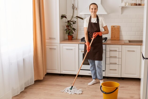 Retrato completo de una atractiva y alegre mujer trabajadora que hace un rápido trabajo doméstico lavando el suelo en una moderna cocina interior blanca y clara con delantal marrón mirando a la cámara