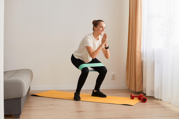 Retrato completo de una atractiva y encantadora mujer deportiva con pantalones blanco y leggins negros, haciendo abdominales con banda elástica, haciendo ejercicio en casa, fitness y estilo de vida saludable.