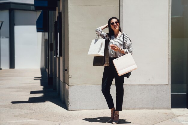 retrato completo de una atractiva joven asiática parada con bolsas de compras en el centro comercial de la ciudad de stanford en un día soleado con gafas de sol. hermosa dama confiada en tacones altos mueve el pelo largo.