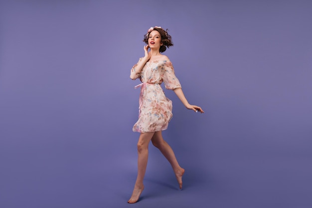 Retrato completo de uma garota inspirada no vestido de verão pulando no estúdio Incrível senhora europeia com flores no cabelo dançando no fundo violeta