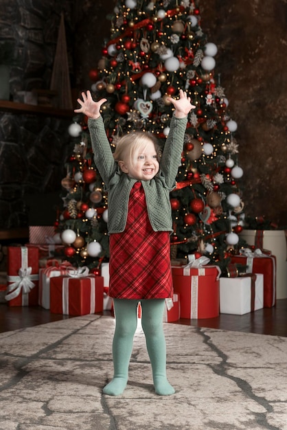 Retrato completo de uma linda garota de três anos com as mãos levantadas perto da árvore de Natal Feliz Natal Ano Novo