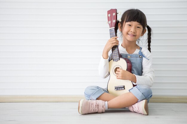 Retrato completo de uma menina bonita tocando ukulele contra uma parede branca em casa