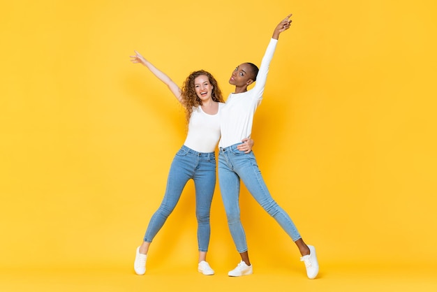 Retrato completo de dos amigas milenarias interraciales sonrientes abrazándose y levantando las manos en un estudio aislado de fondo de color amarillo