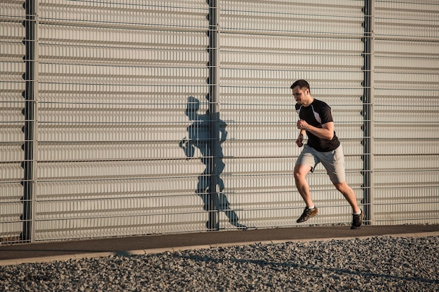 Retrato completo de un hombre atlético corriendo a lo largo de una hermosa pared gris al aire libre, un joven corredor musculoso haciendo ejercicio mientras trotaba en el parque. Atardecer