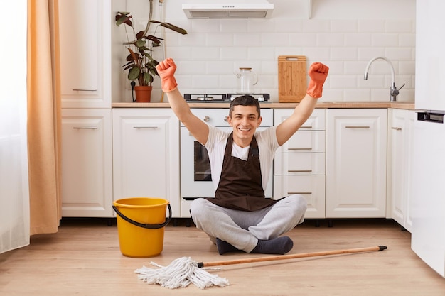 Retrato completo de un hombre feliz y guapo sentado en el suelo en la cocina con los puños apretados feliz de terminar de limpiar la casa mirando la cámara con expresión positiva