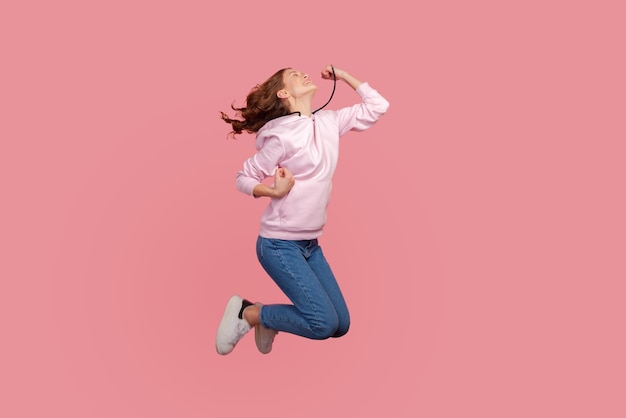 Retrato completo de una joven emocionada con capucha y jeans que gesticula el éxito mientras salta, gana y logra la meta. Disparo de estudio interior aislado sobre fondo rosa