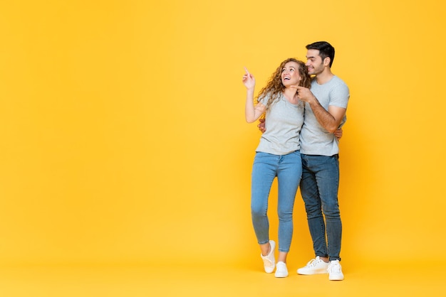 Retrato completo de una joven pareja interracial feliz abrazándose y apuntándose con la mano para copiar el espacio a un lado en un aislado fondo de estudio amarillo