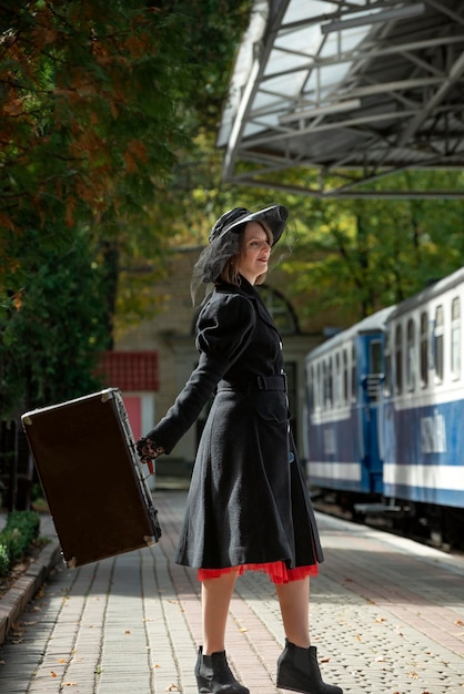 Retrato completo de mujer con abrigo vintage y sombrero con maleta retro esperando el tren en la plataforma de la estación