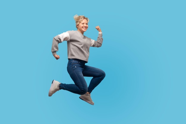 Retrato completo de mujer con cabello corto en sudadera y jeans saltando o corriendo en el aire luciendo enérgico y alegre corriendo para los fines de semana en estudio interior aislado en fondo azul