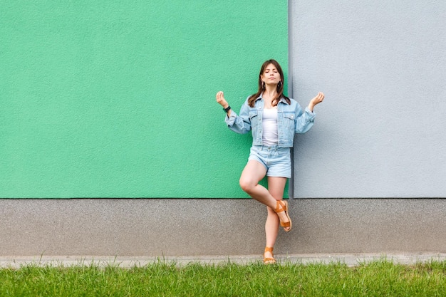 Retrato completo de una mujer hermosa con jeans casuales estilo denim en verano parada cerca de una pared verde y azul claro y haciendo yoga meditando con los ojos cerrados y los brazos levantados.