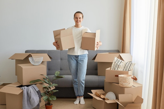 Retrato completo de una mujer positiva y satisfecha con una camiseta blanca de pie con cajas de cartón que se mudan a una casa nueva que desempaca una pila personal que sostiene paquetes el día de la mudanza