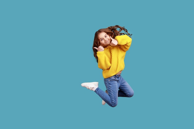 Retrato completo de una niña feliz y encantadora saltando alto y mostrando el pulgar hacia la cámara, usando un suéter amarillo de estilo informal. Disparo de estudio interior aislado sobre fondo azul.