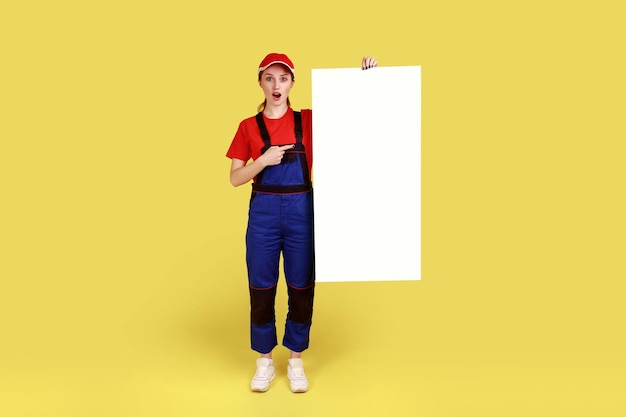 Retrato completo de una trabajadora asombrada sosteniendo y señalando un cartel de papel blanco con un espacio vacío para publicidad o texto promocional Foto de estudio en interiores aislada en fondo amarillo
