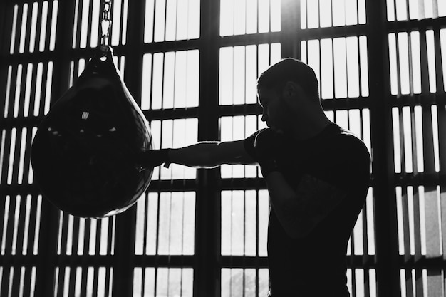 Retrato conceptual de un brutal boxeador tatuado que entrena en el ring y golpea un saco de boxeo