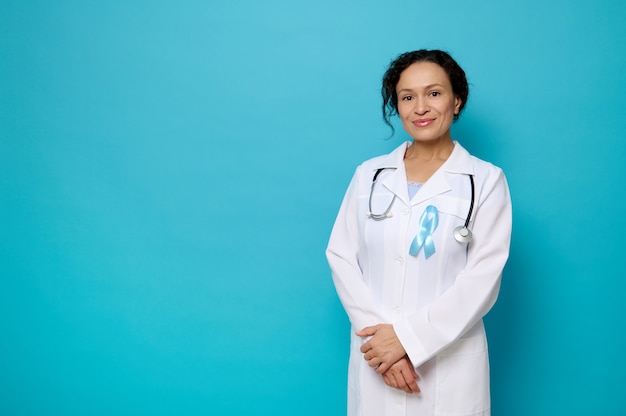 Retrato confiante da médica em um vestido branco médico com fita azul da consciência, símbolo do dia mundial da diabetes, sorrisos, olhando para a câmera posando contra um fundo de cor azul com espaço de cópia