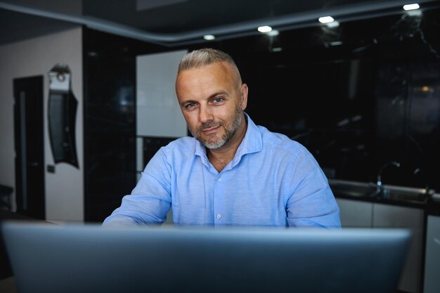 Retrato confiante de um charmoso homem europeu de meia-idade, sentado em frente a um laptop e sorrindo, olhando para a câmera contra um fundo de cozinha elegante em casa