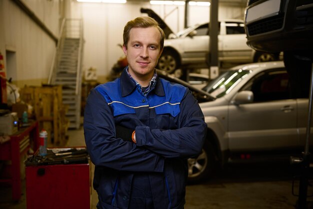 Retrato confiante de um mecânico de automóveis vestido com uniforme profissional, olhando para a câmera em pé com os braços cruzados na oficina de automóveis. Reparação automóvel, diagnóstico e serviço profissional no serviço automóvel