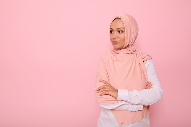 Retrato confiante de uma bela mulher árabe muçulmana com olhar atraente e cabeça coberta com hijab rosa, fica três quartos do fundo colorido com espaço de cópia e desvia o olhar para o espaço de cópia