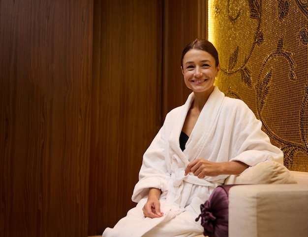 Retrato confiante de uma mulher bonita caucasiana de meia-idade relaxada e serena em um roupão de banho branco, sentada em um sofá, com um sorriso dentuço olhando para a câmera enquanto descansa em uma luxuosa área de salão de spa