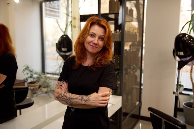 Retrato confiante de uma ruiva na moda, uma mulher bonita cabeleireiro com braços tatuados, sorrisos, olhando para a câmera posando com os braços cruzados contra um espelho em um salão de beleza