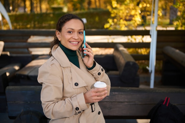 Retrato confiante na altura da cintura de uma jovem alegre falando no celular, segurando um copo de papel para viagem com café quente e sorrisos, olhando para a câmera, enquanto relaxa na cafeteria do bosque de carvalho