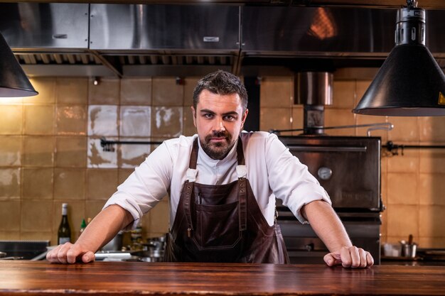 Retrato de confianza guapo chef barbudo en delantal de cuero de pie en el mostrador de madera en la cocina abierta