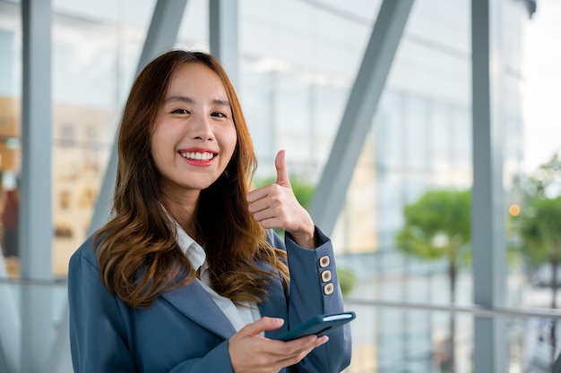 Retrato de confianza mujer de negocios asiática sonriendo y mirando a la cámara y sosteniendo su teléfono celular Ella levanta el pulgar al dedo siguiente espejo escena de construcción en el fondo en la oficina