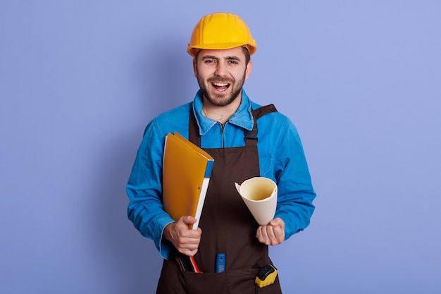 Retrato del constructor hermoso emocional que se coloca aislado sobre la pared azul en el estudio, sosteniendo la caja de papel, abriendo la boca ampliamente, gritando, teniendo barba, vistiendo mono azul, casco y delantal marrón.