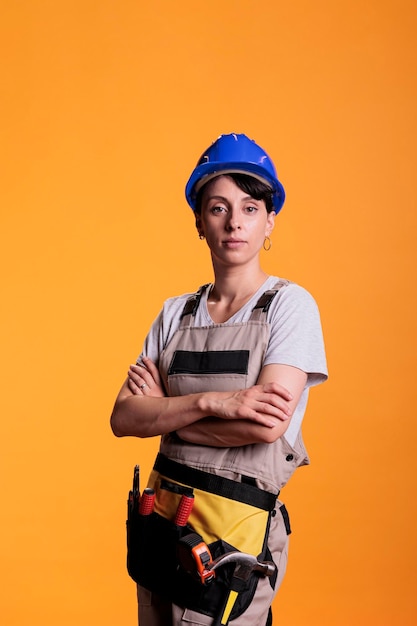 Retrato de un constructor profesional posando con los brazos cruzados en el estudio. Mujer profesional en la industria de la construcción que parece segura, parada en overoles de renovación sobre fondo amarillo.