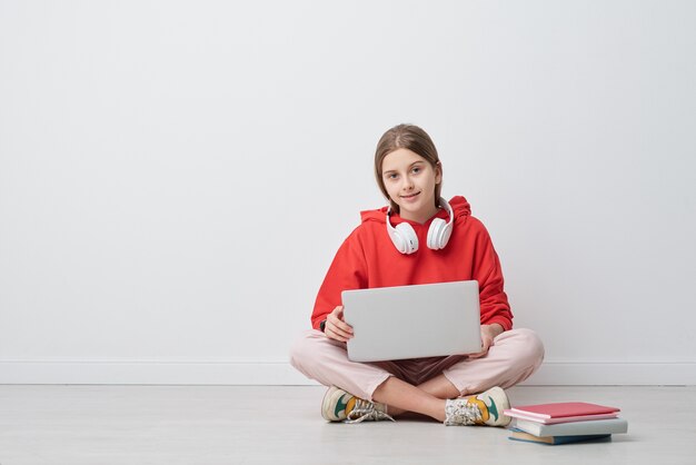 Retrato de contenido chica de secundaria moderna en sudadera con capucha roja sentada con las piernas cruzadas en el piso y usando una computadora portátil