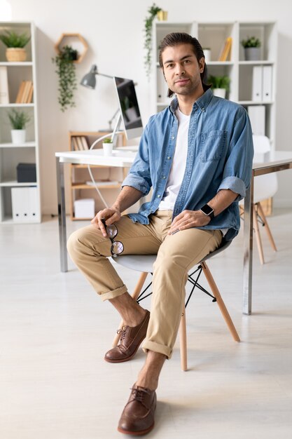 Retrato de contenido guapo joven morena en camisa de mezclilla sentado en una silla y sosteniendo anteojos en la oficina moderna