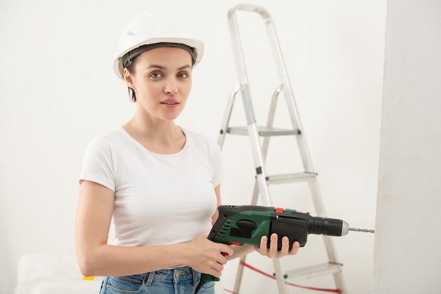 Retrato de contenido joven en casco con taladro eléctrico mientras realiza la renovación en piso nuevo