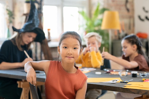 Retrato de contenido niña asiática sentada contra el maestro y los niños en la clase de manualidades de Halloween