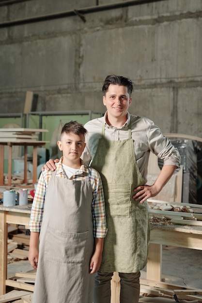 Retrato de contenido padre de mediana edad en delantal sosteniendo la mano sobre el hombro del hijo adolescente en el taller de muebles