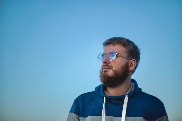 Retrato contra el cielo de un joven con gafas con barba al atardecer en un cálido día de verano