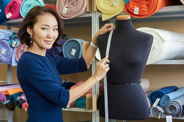 Retrato de una costurera asiática sonriente tomando medidas de un maniquí con un centímetro