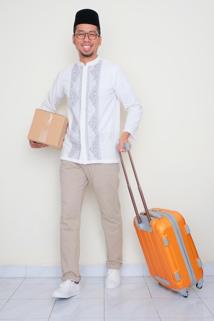 Retrato de cuerpo completo de un hombre musulmán tirando de una maleta y llevando cartón con cara feliz