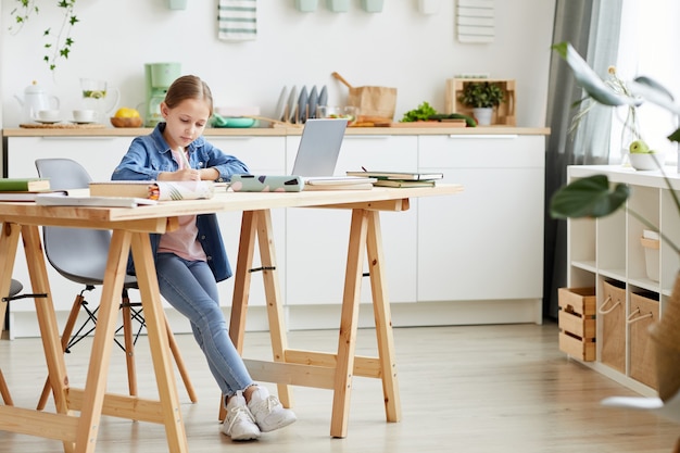 Retrato de cuerpo entero de una adolescente rubia escribiendo en un cuaderno mientras hace la tarea o estudia en casa en un interior acogedor, espacio de copia