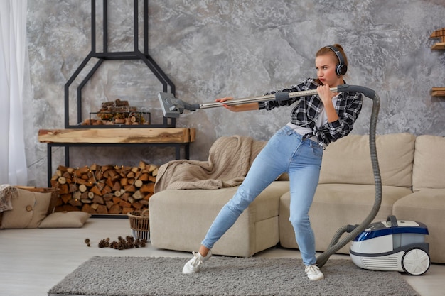 Retrato de cuerpo entero de una alegre joven de 20 años escuchando música a través de auriculares y divirtiéndose con la aspiradora en la casa.