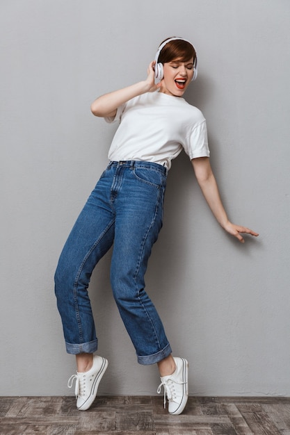 Retrato de cuerpo entero de alegre joven sonriendo y escuchando música con auriculares aislado sobre pared gris